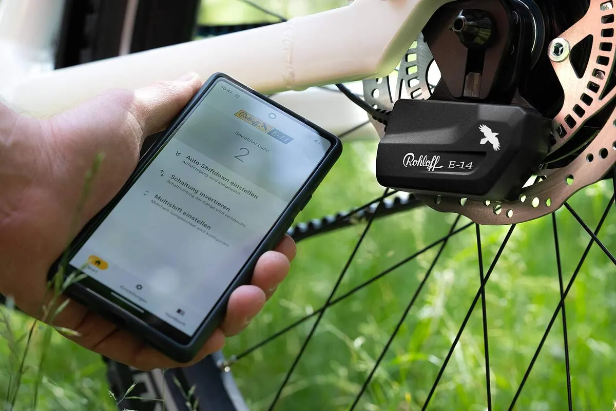 Ein Telefon mit der Startseite der Rohloff-E-14-2.0-App neben der Schalteinheit am Hinterrad